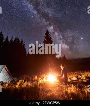 Side view of female tourist having a rest by campfire on background of tent and spruce forest under starry sky on which milky way is visible. Bonfire with a big flame under the night sky Stock Photo
