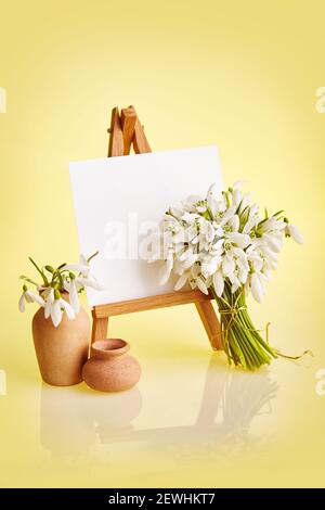 Bouquet of Galanthus flowers and a small easel with a white paper a on a yellow background. Greeting card with a copy space Stock Photo