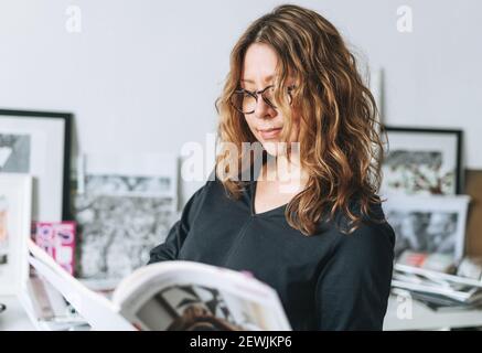 Charming smiling adult woman middle aged woman with curly hair architect designer with magazine catalog in hands in the modern office Stock Photo