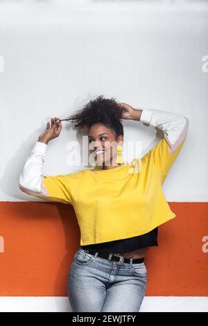 Positive African American millennial female in trendy yellow shirt touching curly hair and looking away while dancing near wall.