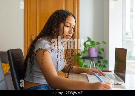 girl writing some notes on a piece of paper sitting at the living room table while she's working the computer from home. Stock Photo