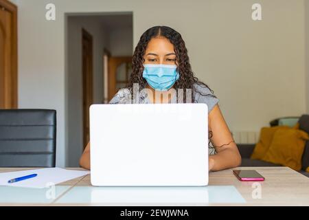 young woman with a mask on her face looking at her cell phone with the computer on the living room table Stock Photo