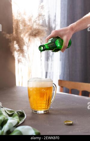 A man pours light beer into a glass from a bottle, a full glass mug of beer with foam on a gray table against the background of a window Stock Photo