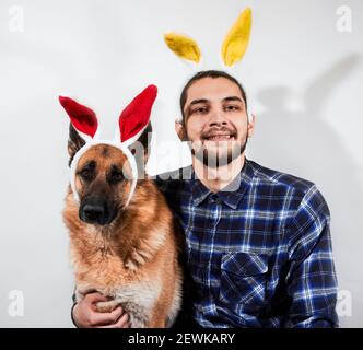 Caucasian man with beard and yellow rabbit ears hugs German shepherd with red hare ears and smiles broadly. Creative banner for Easter with man and hi Stock Photo