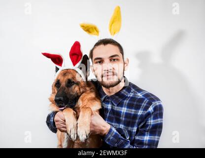 Young handsome Caucasian man with beard hugs his dog in rabbit costume. Creative banner with man and German shepherd with Easter bunny ears on white b Stock Photo