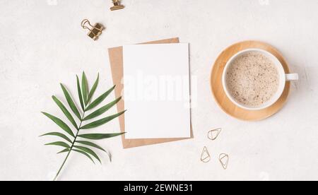 Craft paper envelope with white blank paper note mockup with cup of coffee, gold binder clips and green leaf on white background. Flat lay, top view. Stock Photo