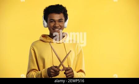 cheerful african american teenager in wireless headphones holding drumsticks isolated on yellow Stock Photo