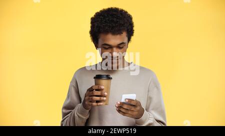 happy african american teenager in sweatshirt holding coffee to go and using smartphone isolated on yellow Stock Photo