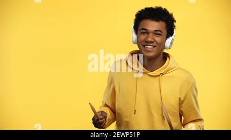happy african american teenager in wireless headphones holding drumsticks isolated on yellow Stock Photo