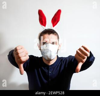Man with ears of Easter bunny. Caucasian man with red hare ears wears medical protective mask and puts dislike with both hands. Celebrate Easter in se Stock Photo