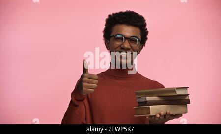 cheerful african american teenage boy in glasses holding books while showing thumb up isolated on pink Stock Photo