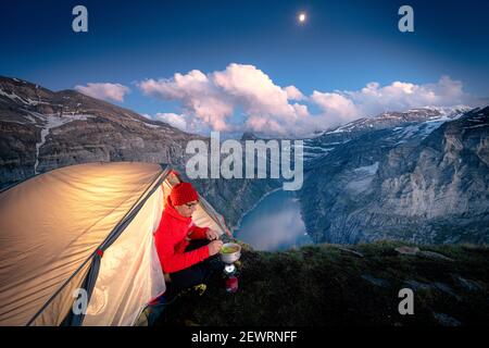 Hiker man with tent using camping stove on ridge above lake Limmernsee, Canton of Glarus, Switzerland, Europe Stock Photo