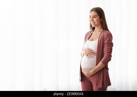 Joyful Pregnant Woman Embracing Her Belly While Standing Near Window At Home Stock Photo