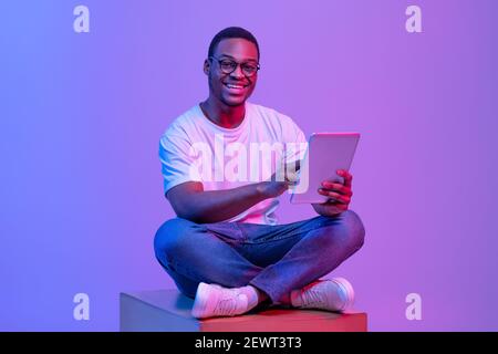 Black Student Guy With Digital Tablet Sitting On Cube Under Neon Light Stock Photo