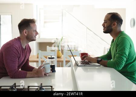 Multi ethnic gay male couple smiling and sitting in kitchen drinking coffee and using laptop at home Stock Photo