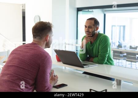 Multi ethnic gay male couple smiling, sitting in kitchen drinking coffee and using laptop at home Stock Photo