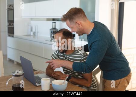 Multi ethnic gay male couple smiling and eating breakfast and using laptop at home Stock Photo