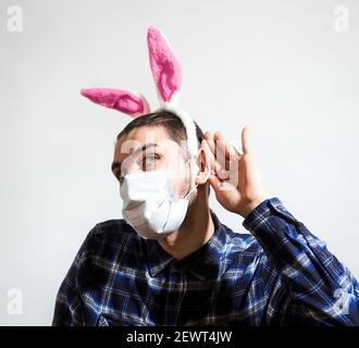 Charming young Caucasian man wearing medical protective mask and pink rabbit ears listens intently. Creative Easter background for your ads and promot Stock Photo