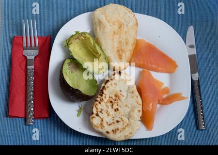 two naan breads toasted with salmon and half avocado on a white plate , red napkin, fork and knife , on blue table mat Stock Photo