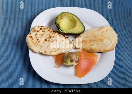 two naan breads toasted with salmon and half avocado on a white plate on blue table mat Stock Photo