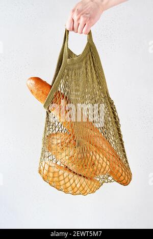 Woman hand holding string shopping or cotton shopper reusable mesh bag with bread before gray background. Zero waste concept. Space for text. Mock up. Stock Photo