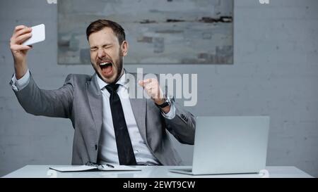 excited businessman with open mouth taking selfie on smartphone near laptop on desk Stock Photo