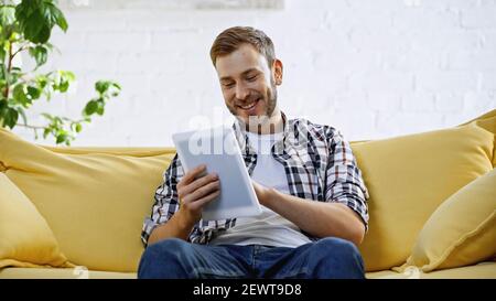 cheerful and bearded man using digital tablet while sitting on couch Stock Photo