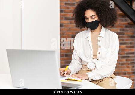 African American female student or businesswoman with curly hair wearing black protective face mask sitting in the coffee shop or library, doing research, looking at laptop, taking notes to notebook Stock Photo