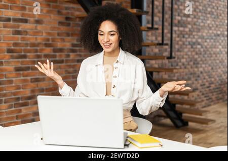 Confused and surprised young stylish African American female freelancer businesswoman or student sitting at the desk with laptop, video calling and gesturing, wondering and asking what question Stock Photo