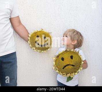 Toddler boy chooses the mood with the help of his dad. Two large sunflower heads with funny and sad faces painted on them. Childhood, parenting. Choic Stock Photo