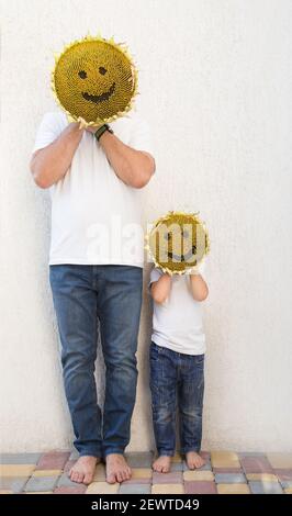 man and boy in white t-shirt and jeans. holding yellow sunflower head in front of faces, on which muzzle with smile is drawn. concept of positive, opt Stock Photo