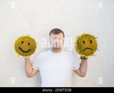 against light background, disgruntled man holds in his hands two large sunflower heads with painted cheerful and sad faces on them. concept of choosin