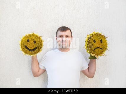 satisfied man holds in his hands two large sunflower heads with painted cheerful and sad faces on them. the concept of choosing a mood, life position. Stock Photo