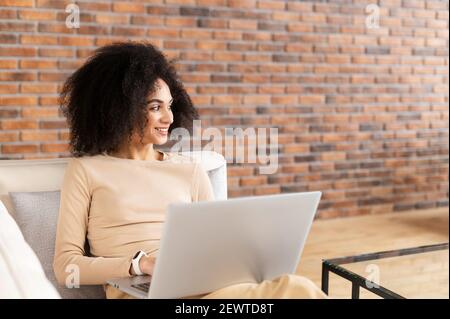 Young African American female freelancer with Afro hairstyle sitting on the couch and working on the project, studying, blogging, resting and chatting online, planning daydreaming, and looking away Stock Photo