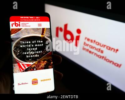 Person holding mobile phone with web page of food company Restaurant Brands International (RBI) on screen in front of logo. Focus on phone display. Stock Photo