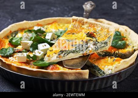 feta spinach cheddar cheese quiche in a baking dish on a wooden board on a grey concrete table with a slice on a spatula, horizontal view from above, Stock Photo