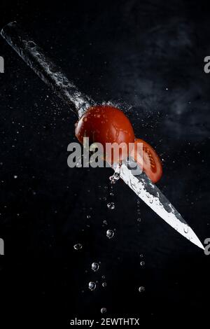 Fresh tomato cutting with a knife and flying in motion on the black background with splashes. Stock Photo