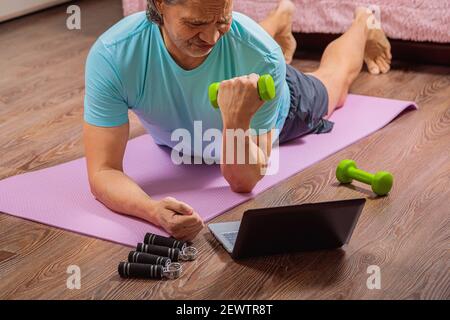 50-year-old man performs exercises while lying on the rug at home, looking at the computer. During a pandemic, a person trains in an apartment via the Stock Photo
