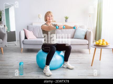 Lovely mature woman sitting on fitness ball, working out with dumbbells in her home gym, empty space. Active lifesyle Stock Photo