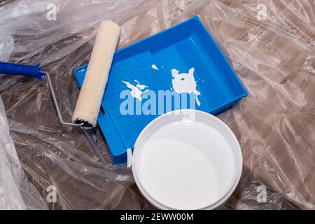 Tools for priming plastered surfaces for further painting.Paint roller in a blue tray with white paint on dirty floor background. Renovation, painting Stock Photo