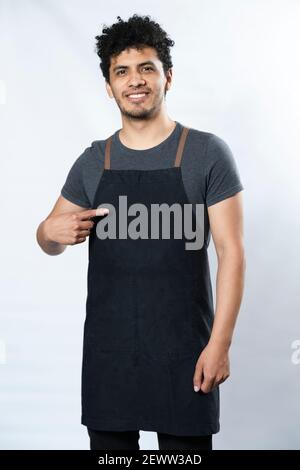 Black apron mockup - Hispanic young man pointing at apron smiling on white background - young worker - template for text on clothes Stock Photo