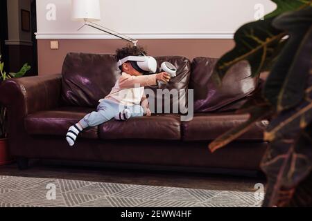 Little boy wearing VR glasses while sitting on sofa. Kid with virtual reality goggles and controller playing a video game Stock Photo