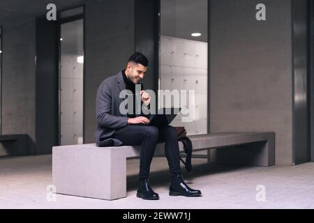 Businessman with glasses smiling and sitting on a bench typing on his laptop. Stock Photo