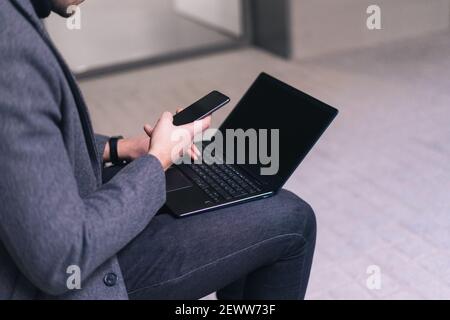 Unrecognizable man with his cell phone while sitting on a bench with his laptop with black screen. Stock Photo