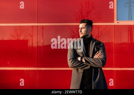 Young businessman with backpack and arms crossed in front of a red wall at sunset. Stock Photo