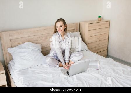 Photo of young joyful woman in pajama typing on laptop and smiling while sitting on bed in light room Stock Photo