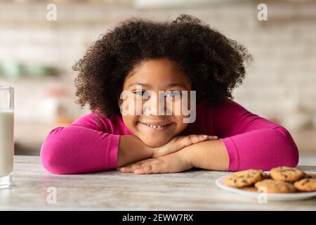 Adorable Little Black Girl Sitting At Table With Cookies And Milk Stock Photo