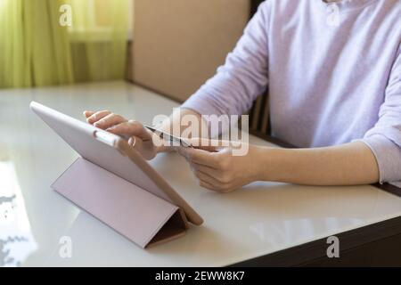 Young caucasian unrecognizable woman makes purchases in online store, uses tablet and credit card, sits at table. Online shopping concept, stay home, Stock Photo