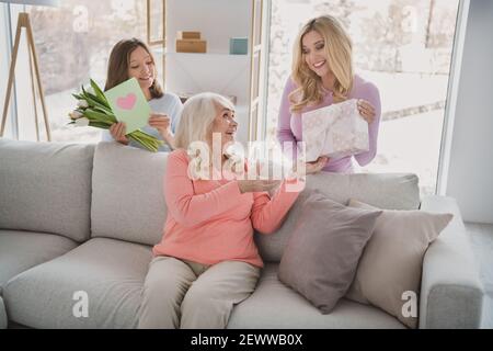 Photo portrait of grandmother receiving gift box postcard flowers on birthday from daughter granddaughter sitting on sofa Stock Photo