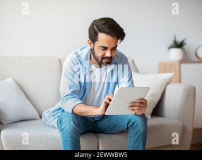Handsome Eastern Guy Using Digital Tablet While Relaxing On Couch At Home Stock Photo